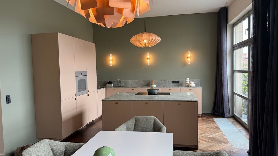 Modern kitchen-dining area with muted green walls, pale built-in cabinets, and a large marble-topped island with an integrated cooktop. Sculptural pendant lights hang over the room, and a tall window with dark curtains brings in daylight above herringbone wood flooring.