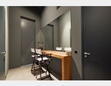 Minimalistisch eingerichteter Raum mit grauen Wänden, einem großen Wandspiegel und einem schlichten Holztisch. Zwei klappbare Stühle mit weißen Sitzbezügen stehen vor dem Tisch, der als Schmink- oder Arbeitsbereich genutzt werden könnte.