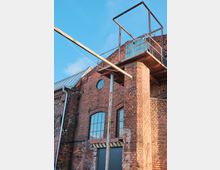 Backsteinfassade eines alten Industriegebäudes mit großen rechteckigen Fenstern und einem runden Oberlicht. Ein rostiger Metallpfosten und eine erhöhte Plattform mit Geländer aus Stahl sowie einer schmalen Überdachung heben sich vor dem blauen Himmel ab.