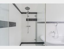 Modernes Badezimmer mit bodengleicher Dusche hinter Glaswand, weiß gefliest und mit dunklem Fliesenband akzentuiert. Rechts ist eine freistehende Eckbadewanne mit Handbrause und Armatur zu sehen.