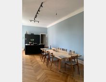 Wohnraum mit hellblauer Wand und Fischgrätenparkettboden. Im Vordergrund ein großer Holztisch mit Holzstühlen, im Hintergrund eine schwarze Küchenzeile mit Dekofigur und einer offenen Tür. An der Decke eine schwarze Schienenbeleuchtung und eine Einzelbirne.