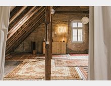 Dachgeschossraum mit sichtbaren Holzbalken, rustikalen Wänden und Holzboden, dekoriert mit mehreren orientalischen Teppichen. Im Hintergrund stehen ein kleines Holzregal, eine Tischlampe mit der leuchtenden Aufschrift „OM“ und ein Fenster mit weißem Rahmen.