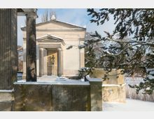 Schneebedeckter Eingang zu einem klassizistischen Gebäude mit Säulen und einem Dreiecksgiebel. Im Inneren des Eingangsbereichs steht eine Büste auf einem Sockel, umgeben von winterlicher Vegetation und einer Steinmauer.