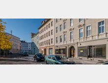 Straßenszene mit klassischen Wohn- und Geschäftshäusern im Altbaustil, gekennzeichnet durch verzierte Fassaden und hohe Fenster. Parkende Autos säumen die gepflasterte Straße, während ein Baum mit herbstlich gelbem Laub links im Bild sichtbar ist.