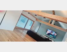 Modernes Wohnzimmer mit sichtbaren Holzbalken an der Decke, einem großen Fenster mit Jalousien, Holzboden, einer schwarzen Couch und einem an der Wand befestigten Flachbildfernseher.