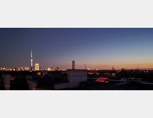 Skyline von Berlin bei Dämmerung mit Blick auf den Fernsehturm und beleuchtete Gebäude im Hintergrund. Im Vordergrund sind die Dächer von Wohnhäusern sichtbar, teils mit Schornsteinen und einem rot beleuchteten Dachfenster.