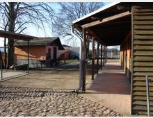 Hof mit rustikaler Architektur: Im Vordergrund ist ein gepflasterter Weg zu sehen, rechts ein langes Gebäude mit Holzverkleidung und Überdachung, links ein orangefarbenes Haus mit Satteldach und kleinen Pflanzen. Im Hintergrund sind Bäume und weiteres ländliches Gelände erkennbar.
