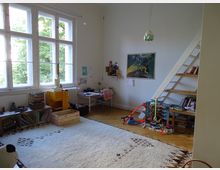 Ein Kinderzimmer mit großen Fenstern, die viel Tageslicht hereinlassen, einem hellen Teppich, Regalen mit Büchern und Spielzeug sowie einer kleinen Holzleiter, die zu einem erhöhten Bereich führt. Ein Schreibtisch befindet sich an der Wand, daneben ist eine Spielzeug-Autorennbahn aufgebaut, und an der Wand hängt ein gerahmtes Poster.