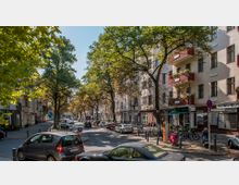 Straßenszene in einer Wohngegend mit mehrstöckigen Altbauhäusern, vielen Bäumen entlang der Straße und parkenden Autos. Im Erdgeschoss der Gebäude befinden sich Geschäfte, Cafés und Balkone mit Blumen.