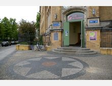 Backsteingebäude mit breitem Eingang, dessen grüne Tür offensteht und über eine Treppe erreichbar ist. Davor ein gepflasterter Platz mit einem sternförmigen Mosaik und mehrere abgestellte Fahrräder; links daneben ein schwarzes schmiedeeisernes Geländer und eine Baumallee entlang der Straße.