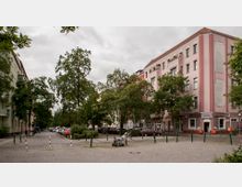 Ansicht eines städtischen Platzes mit Kopfsteinpflaster, umgeben von mehrstöckigen Wohngebäuden, darunter ein rosa Gebäude mit einem Friseurgeschäft im Erdgeschoss. Im Vordergrund stehen einzelne Bäume, Laternen und ein Kinderwagen, im Hintergrund sind parkende Autos zu sehen.