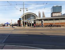 Eine Straßenansicht mit dem Eingang zur U-Bahn-Station "Warschauer Straße" in einem Gebäude mit einem großen, gewölbten Glasportal. Vor der Station befindet sich ein Fahrradständer mit zahlreichen Fahrrädern, daneben eine Straße mit Straßenbahn- und Fahrbahnschienen, und im Hintergrund stehen moderne Bürogebäude.