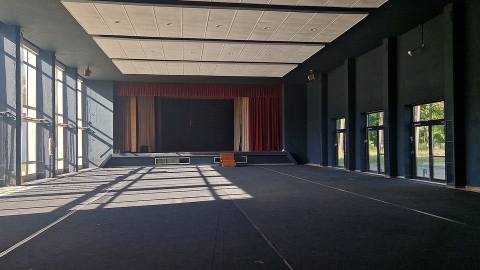 Großer, dunkler Veranstaltungssaal mit einer Bühne im Hintergrund, umrahmt von roten Vorhängen. Links befinden sich hohe Fenster, die Sonnenlicht in den Raum lassen, und rechts sind mehrere verglaste Türen sichtbar.