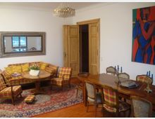 Wohnzimmer mit klassischer Einrichtung, bestehend aus einem runden Holztisch, einer karierten Sitzgarnitur und einem großen rechteckigen Esstisch mit passenden Stühlen. An der Wand hängt ein großer Spiegel, daneben ein buntes Gemälde, und ein Kronleuchter hängt von der Decke; der Boden ist mit einem Orientteppich bedeckt.