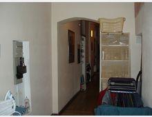 Ein schmaler Flur in einem Wohnbereich mit hellem Anstrich, einem gewölbten Durchgang und Holzboden. Im Vordergrund stehen ein Wäscheständer und ein Regal mit geflochtenen Körben, an den Wänden hängen Bilder.