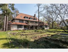 Großes, zweistöckiges Landhaus mit rotem Ziegeldach und mehreren Gauben, umgeben von einem weitläufigen Garten mit Bäumen und Beeten. Im Vordergrund sind eine terrassenartige Rasenfläche und ein kleiner, überwucherter Teich zu sehen.