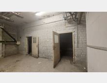 Ein leerer Raum mit abgenutzten weißen Backsteinwänden, zwei geöffneten Türen mit Glasfenstern und dunklen Durchgängen dahinter. Oben sind Leitungen und Rohre sichtbar, der Boden wirkt staubig und unsauber.