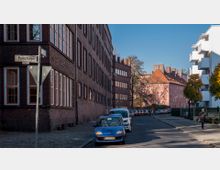 Straßenszene in einem Wohngebiet mit Backsteingebäuden und mehreren parkenden Autos entlang der Straße. Auf der rechten Seite befinden sich moderne weiße Gebäude sowie vereinzelte Bäume, und im Vordergrund ist ein Straßenschild mit der Aufschrift „Parkstraße“ sichtbar.