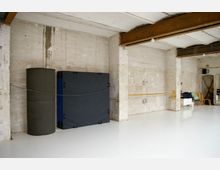 Ein heller Raum mit weißem Boden und rauen, unverputzten Backsteinwänden. An einer Wand befinden sich große Matten und ein zusammengerollter Schaumstoff, daneben eine schmale Ballettstange; im Hintergrund sind Stühle, Regale und eine Tür sichtbar.
