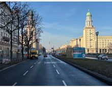 Breite mehrspurige Stadtstraße mit Mittelstreifen und wenig Verkehr, gesäumt von hohen Altbauten und Laternen. Im Hintergrund ragen der Berliner Fernsehturm sowie ein markantes Hochhaus mit grünem Kuppeldach in den blauen Himmel; rechts steht ein großes Werbeplakat am Straßenrand.