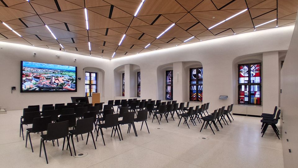 Moderner Veranstaltungs- und Vortragsraum mit Reihen schwarzer Stühle, hellem Boden und weißer Wand mit großen Rundbogenfenstern aus farbigem Glas. An der Wand hängt ein großer Bildschirm, die Decke besteht aus geometrischen Holzpaneelen mit integrierten Lichtleisten.