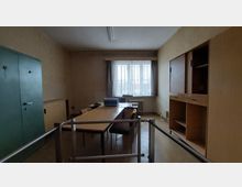 Ein einfach eingerichtetes Büro mit einem rechteckigen Holztisch, mehreren Stühlen, einem grünen Metallschrank auf der linken Seite und einem Holzregal mit Schiebetüren auf der rechten Seite. Das Fenster im Hintergrund ist vergittert und mit Gardinen versehen; eine Schreibmaschine steht auf dem Tisch.