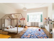 Ein kinderfreundlich eingerichtetes Zimmer mit einem Holzbett in Hausform, einem bunten Teppich und Spielzeug. Im Hintergrund befindet sich eine große Fensterfront mit grünlichen Vorhängen, die viel Licht in den Raum lässt.