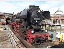 Historische Dampflokomotive mit der Aufschrift „52 8177-9“ steht auf einem Drehscheiben-Gleis vor einem rundbogigen Backstein-Lokschuppen. Im Hintergrund sind weitere Schienen und Gebäude eines Eisenbahnmuseums zu sehen.