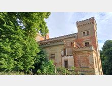 Eine verfallene Backsteinvilla mit Zinnen und einem Eckturm, umgeben von dichter Vegetation und Bäumen. Teile des Gebäudes zeigen beschädigte oder fehlende Fenster und bröckelnde Fassaden.