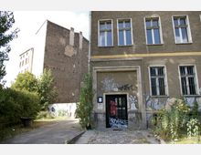 Außenansicht eines alten Gebäudes mit bröckelnder Fassade, mehreren großen Fenstern und Graffiti an den Wänden. Der Eingangsbereich ist mit Schmierereien bedeckt, daneben ist ein mit Pflanzen bewachsener Weg, der zu einem angrenzenden, teils überwucherten Hinterhof führt.