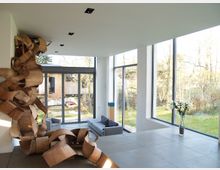 Modernes Wohnzimmer mit großen bodentiefen Fenstern, die den Blick auf einen Garten mit Bäumen freigeben. An der linken Wand befindet sich eine große Holzskulptur, daneben stehen graue Sofas und ein Glastisch mit einer Vase voller Blumen.