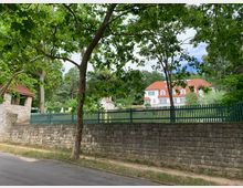 Straßenansicht eines historischen Anwesens mit weißem Gebäude und rotem Ziegeldach, umgeben von einem Zaun und einer Steinmauer. Im Vordergrund stehen grüne Bäume, die teilweise den Blick auf das Gebäude verdecken.