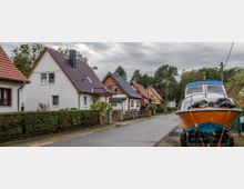 Straße in einem Wohngebiet mit Einfamilienhäusern, die kleine Gärten und Satteldächer haben. Am rechten Straßenrand steht ein orange-blaues Boot auf einem Anhänger, umgeben von Bäumen und bewölktem Himmel.