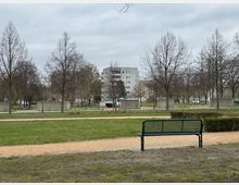 Ein Park mit einer Rasenfläche, mehreren kahlen Bäumen und einer Sitzbank im Vordergrund. Im Hintergrund sind Wege, Betonmauern und ein mehrstöckiges Wohngebäude zu sehen, unter bedecktem Himmel.