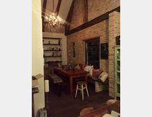 Gemütlicher Wohnbereich mit rustikalem Stil, featuring einer Ziegelsteinwand, sichtbaren Holzbalken und einem funkelnden Kronleuchter. Ein Holztisch mit Bänken und Kissen steht im Vordergrund, daneben ein offenes Regal mit Büchern.