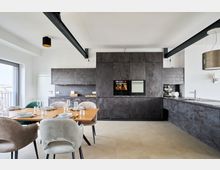 Moderne offene Küche mit dunklen, strukturierten Einbauschränken und integriertem Backofen. Im Vordergrund ein Esstisch aus Holz mit gedeckten Plätzen und Polsterstühlen, daneben eine Arbeitsfläche mit Spüle und Hängelampe.