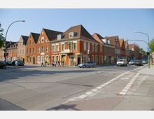 Straßenszene mit einer Kreuzung in einer Stadt; am Straßenrand stehen Backsteingebäude mit spitzen Giebeln und Geschäften, darunter eine Apotheke. Auf der Straße fahren Autos, und Fußgänger überqueren den Zebrastreifen bei Sonnenschein.