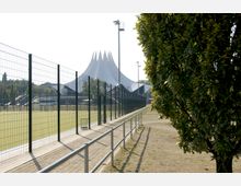 Fußweg entlang eines Zauns, der ein Sportfeld umgibt, im Hintergrund eine markante, weiße Zeltstruktur mit spitzen, geschwungenen Dächern, daneben ein hoher Flutlichtmast und Bäume. Im Vordergrund steht ein Baum, der teilweise den Blick auf das Gelände verdeckt.