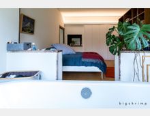 Modernes Schlafzimmer mit minimalistischem Design, bestehend aus einem Bett mit blauer Decke und rotem Überwurf. Im Vordergrund befindet sich der Rand einer Badewanne, links ein Wäschekorb und rechts eine große Monstera-Pflanze auf einer Ablage; im Hintergrund sind weiße Einbauschränke und ein Bild an der Wand zu sehen.