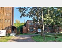 Backstein-Gebäude einer ehemaligen Industrieanlage mit großen rechteckigen Fenstern, flankiert von Bäumen und Efeu-bewachsenen Mauern. Im Vordergrund führt ein gepflasterter Weg mit Schienen durch eine Grünfläche.