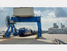 Eine blaue Diesellokomotive steht vor einem großen blauen Hafenkran auf einem Industriegelände. Im Hintergrund sind Industriegebäude, Schienen und ein Schild mit der Aufschrift „Schwedter Hafen“ sichtbar, während Rauch aus einem Schornstein in den Himmel steigt.