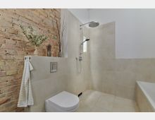 Modern gestaltetes Badezimmer mit freistehender Toilette, einer bodengleichen Dusche mit Regenbrause und hellen Wand- und Bodenfliesen. Auf einer halbhohen Trennwand aus Fliesen vor einer Backsteinwand befinden sich Dekorationselemente wie eine Vase mit Zweigen.