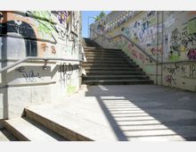 Außenbereich mit einer Treppe aus Beton, flankiert von Wänden mit zahlreichen Graffiti und Geländern aus Metall. Die Szene ist von Sonnenlicht durchzogen, das Schattenstreifen auf den Boden wirft.