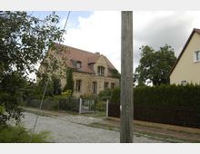 Ein zweigeschossiges Haus aus hellem Backstein mit rotem Ziegeldach, umgeben von einem Garten mit Blumen und Sträuchern, eingefasst von einem Metallzaun. Im Vordergrund verlaufen eine gepflasterte Straße und ein Holzstrommast.