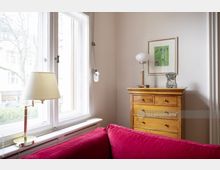 Wohnzimmer-Ecke mit einem großen Fenster, rotem Sofa im Vordergrund, einer Holzkommode mit dekorativen Gegenständen und einer Wandlampe sowie einem gerahmten Kunstwerk an der Wand. Draußen sind Bäume und Gebäude schemenhaft sichtbar.