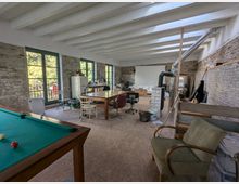 Großer Raum mit rustikaler Einrichtung, sichtbaren Backsteinwänden und weißen Holzbalken an der Decke. Der Raum enthält ein Billardtisch, Holzmöbel, eine Sitzgruppe, einen modernen Kaminofen und breite Fenster, die viel Tageslicht hereinlassen.