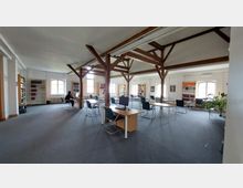 Großer, heller Lesesaal einer Bibliothek mit Holzbalkendecke, mehreren Arbeitstischen mit Stühlen und Computern sowie Regalen voller Bücher entlang der Wände. Große Fenster lassen Tageslicht in den Raum, und eine Person sitzt an einem Tisch mit einem Laptop.