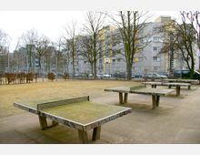 Öffentlicher Platz in einem städtischen Wohngebiet mit mehreren Beton-Tischtennisplatten im Vordergrund, die mit Graffiti bemalt sind. Im Hintergrund sind mehrstöckige Wohnhäuser, geparkte Autos, kahle Bäume und ein eingezäunter Bereich zu sehen.