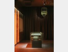 Ein dunkler Raum mit dunklen Vorhängen im Hintergrund, einem rechteckigen Objekt in Gitteroptik im Vordergrund, das ein technisches Gerät umgibt, sowie einer an der Decke montierten Lautsprecheranlage. Links sind beleuchtete orangefarbene Wände zu sehen.