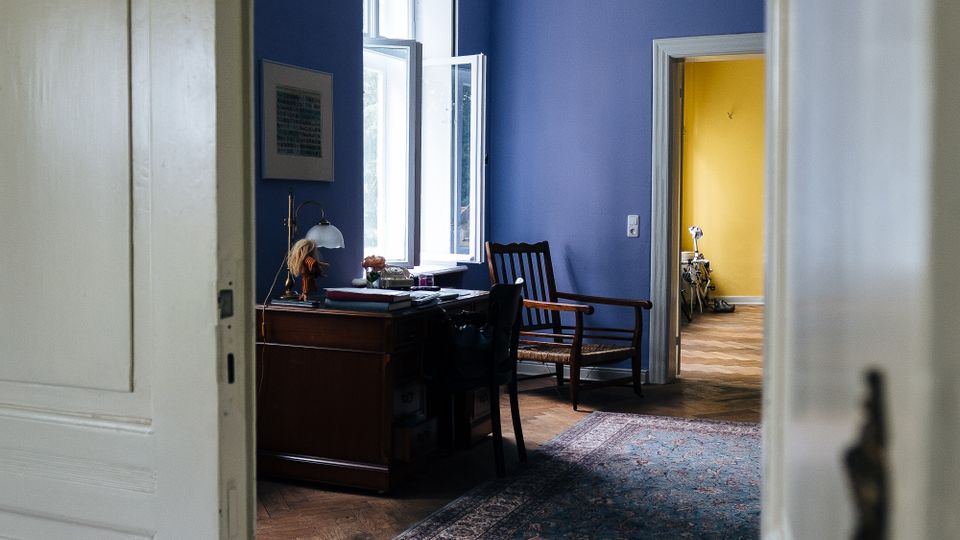 Blick durch eine offene Tür in einen Altbau-Raum mit blau gestrichenen Wänden, Stuckdecke und Fischgrät-Parkett. Am hohen Fenster stehen ein Schreibtisch und ein Stuhl, ein gemusterter Teppich liegt auf dem Boden, und durch eine weitere Tür ist ein gelb gestrichener Nebenraum zu sehen.